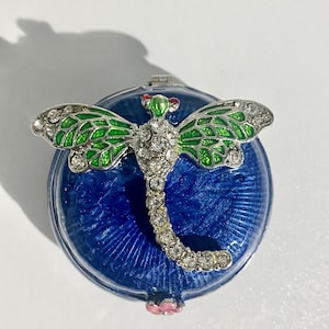 May include: A decorative compact mirror featuring a dragonfly design. The dragonfly has green and silver wings, a body encrusted with clear crystals, and red accents. The compact has a blue, textured background and a pink flower detail.