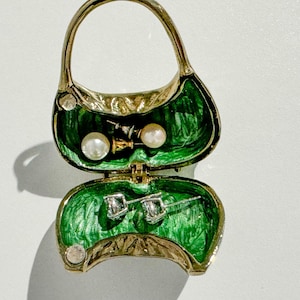 May include: A gold-toned purse-shaped jewelry box with a green interior. The box is open, revealing a pair of pearl earrings and a pair of diamond stud earrings. The purse has a handle and is decorated with small, round embellishments.