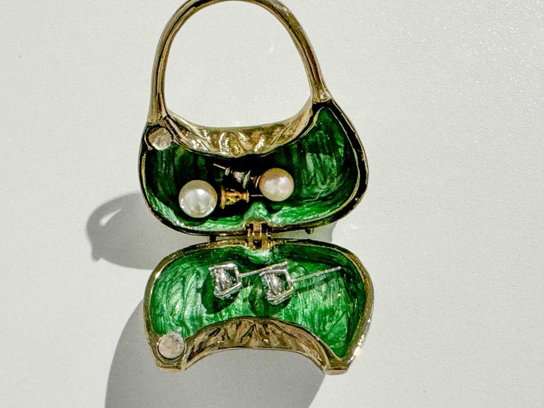 May include: A gold-toned purse-shaped jewelry box with a green interior. The box is open, revealing a pair of pearl earrings and a pair of diamond stud earrings. The purse has a handle and is decorated with small, round embellishments.