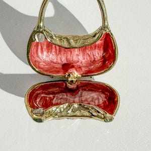 May include: A gold-toned metal purse-shaped trinket box with a hinged opening. The interior is painted with a textured red enamel. The purse has a handle and a clasp to secure the box closed. The overall design is ornate and decorative.
