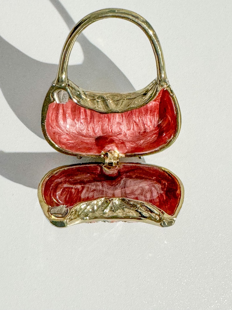 May include: A gold-toned metal purse-shaped trinket box with a hinged opening. The interior is painted with a textured red enamel. The purse has a handle and a clasp to secure the box closed. The overall design is ornate and decorative.