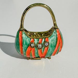 May include: A miniature, decorative handbag with a gold handle and trim. The bag is painted in a green and orange color scheme, with three clear, round gemstones. The handbag is a small, ornate accessory.