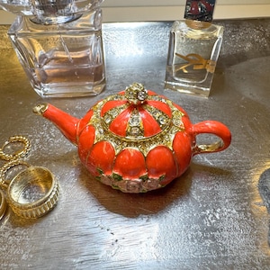 May include: A small, red decorative teapot with gold accents and a jeweled crown on the lid. The teapot is surrounded by perfume bottles and rings on a silver tray. The teapot is a unique decorative item.