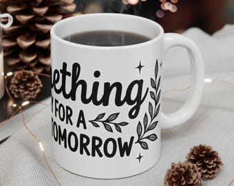 Puede incluir: Taza de cerámica blanca llena de una bebida oscura, con el texto "Something for a Tomorrow" en escritura negra. La taza tiene un asa y está decorada con ilustraciones botánicas. La taza está sobre una superficie gris claro.