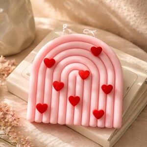 May include: A pink, rainbow-shaped candle decorated with small, red heart-shaped embellishments. The candle is placed on a book, against a neutral backdrop. The candle has a textured surface and is a decorative item.