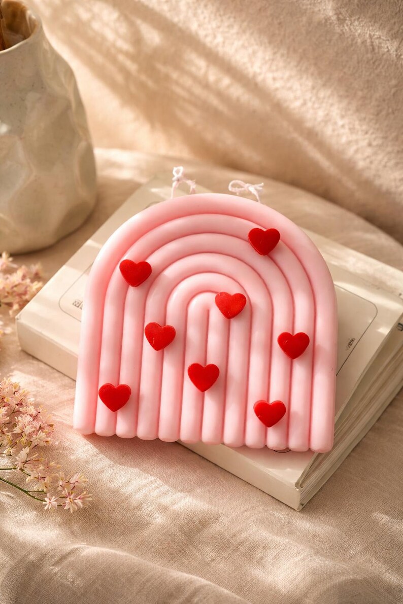 May include: A pink, rainbow-shaped candle decorated with small, red heart-shaped embellishments. The candle is placed on a book, against a neutral backdrop. The candle has a textured surface and is a decorative item.