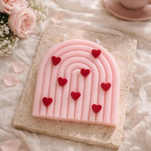 May include: A pink, arch-shaped candle with vertical lines and red heart-shaped decorations. The candle is placed on a rectangular stone base. A cup of tea and flowers are in the background.