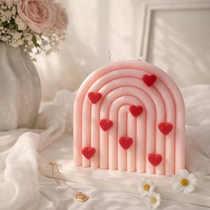 May include: A pink, rainbow-shaped candle decorated with red heart shapes. The candle is on a white surface. The image also features a bouquet of pink roses in a vase and a gold heart pendant. The candle is decorated with nine red hearts.