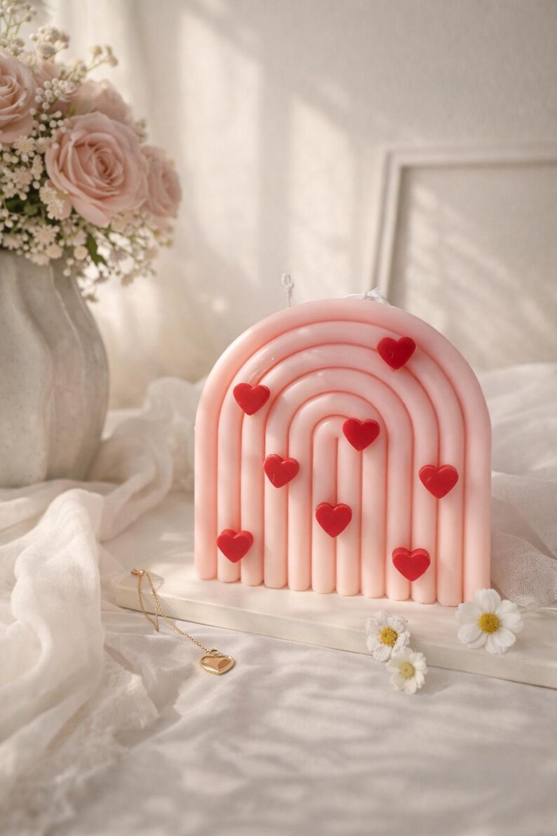 May include: A pink, rainbow-shaped candle decorated with red heart shapes. The candle is on a white surface. The image also features a bouquet of pink roses in a vase and a gold heart pendant. The candle is decorated with nine red hearts.