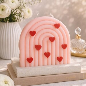May include: A pink, rainbow-shaped candle with red heart accents. The candle is placed on a stack of books, with a vase of white flowers in the background. The candle is a decorative item.