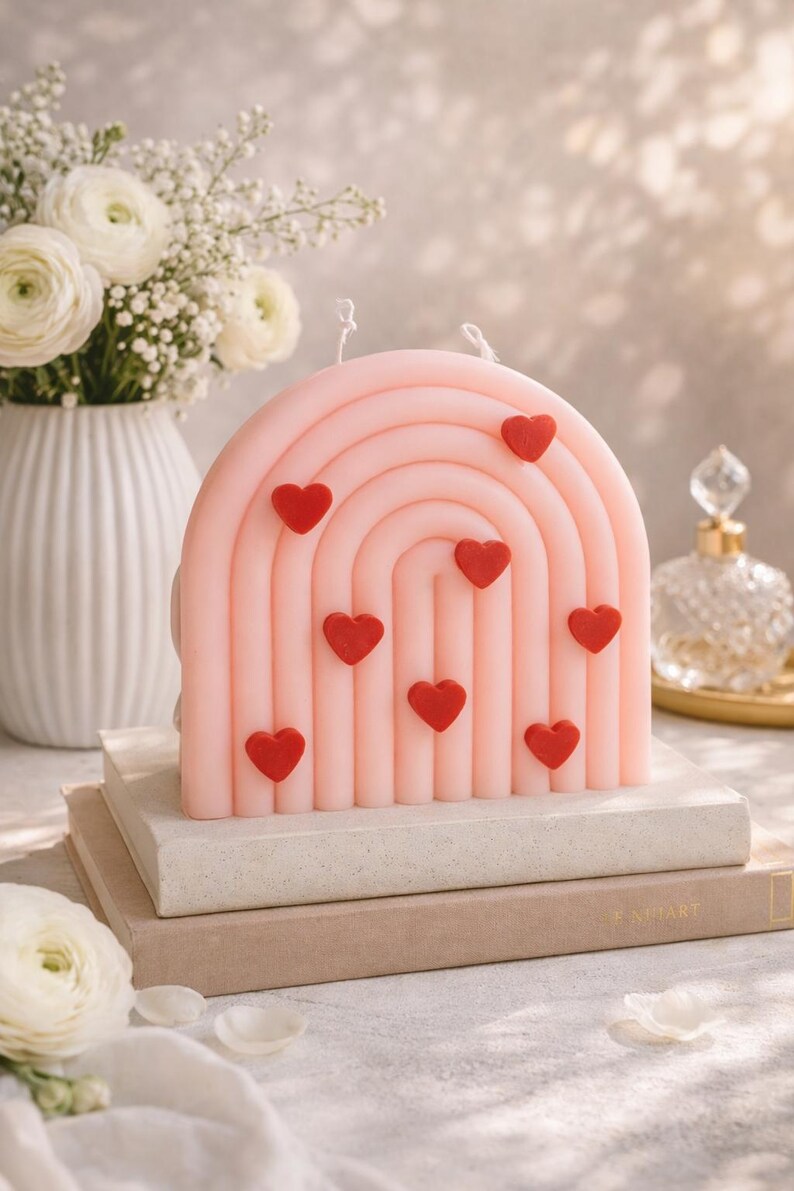 May include: A pink, rainbow-shaped candle with red heart accents. The candle is placed on a stack of books, with a vase of white flowers in the background. The candle is a decorative item.