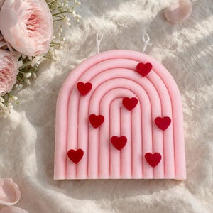 May include: A pink, rainbow-shaped candle with red heart accents. The candle has two white wicks. The candle is a decorative item, perfect for Valentine's Day or as a romantic gift. The candle is surrounded by pink roses and petals.