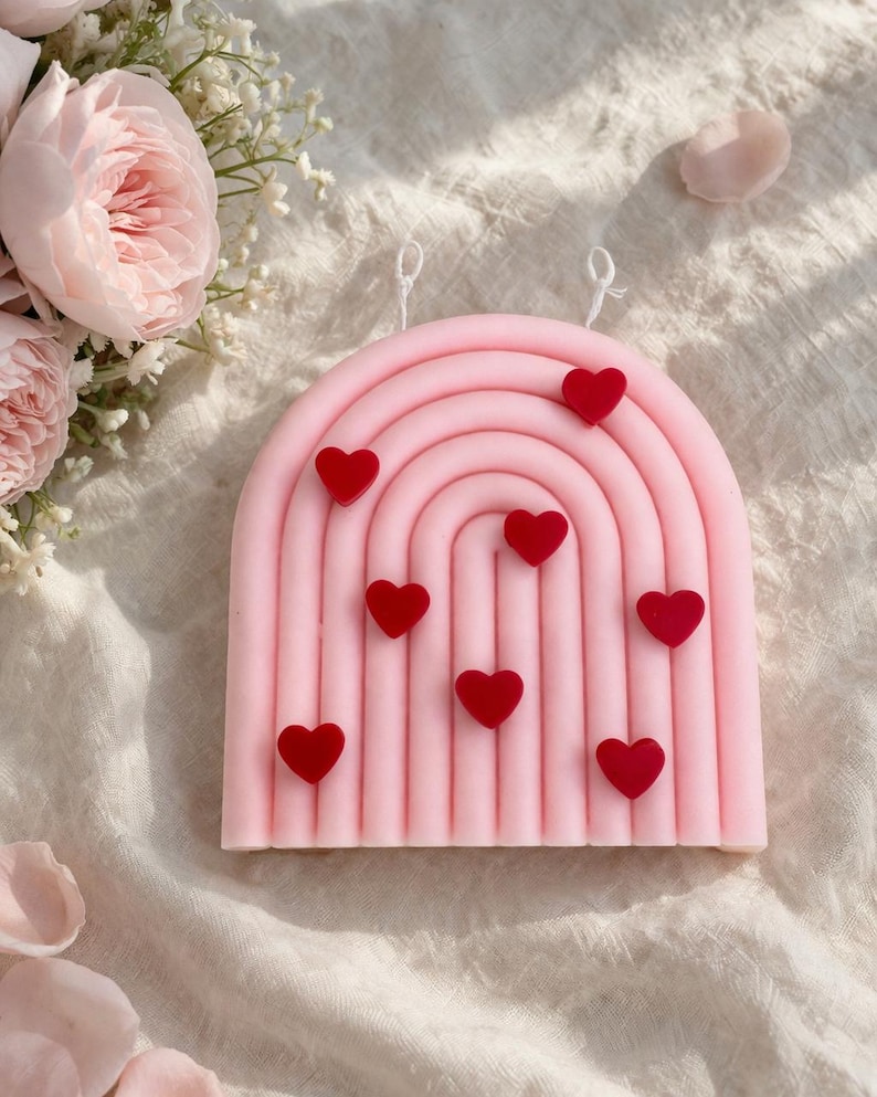 May include: A pink, rainbow-shaped candle with red heart accents. The candle has two white wicks. The candle is a decorative item, perfect for Valentine's Day or as a romantic gift. The candle is surrounded by pink roses and petals.
