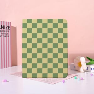 May include: A rectangular, green and tan checkered pattern notebook or tablet cover. The cover is standing upright on a pink surface. The background is a light pink wall. A book with text is visible on the left.