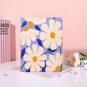 May include: A floral-patterned notebook with a light purple background. Large white daisies with orange centers and blue leaves are arranged across the cover. The notebook is on a white surface with a pink background.