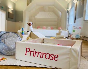 May include: A white inflatable bounce house with a matching ball pit and soft play area. The ball pit is labeled "Primrose" in red lettering. A plush sheep and colorful stacking toys are also visible.