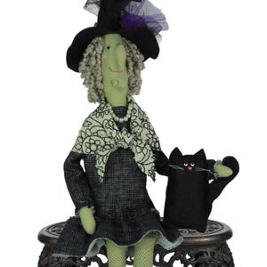 May include: Aunt Ollie & Spritzer doll. The doll is a witch with a green face, gray curly hair, and a black hat. She wears a black dress and sits with a black cat on a decorative wooden stand. The text "It's Sew Relative" is in a circle at the bottom.
