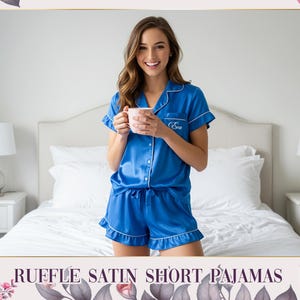 May include: Blue satin short pajamas with white trim. The set includes a short-sleeved button-up top and matching shorts with a ruffle hem. The pajamas have a personalized monogram on the top. The text "RUFFLE SATIN SHORT PAJAMAS" is at the bottom.