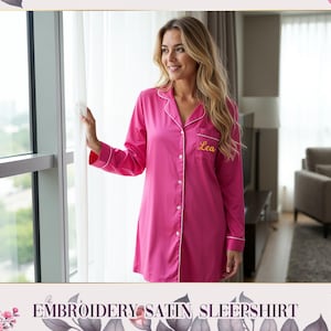 May include: A bright pink button-down sleepshirt featuring long sleeves, a notched collar with white piping, and the name Lea embroidered in gold cursive script on the left chest pocket. The garment has a smooth, satin-like finish.