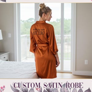 May include: A burnt orange satin robe with the text "Wake Up and Hit Zero" printed on the back. The robe has a matching belt and is knee-length. The image also includes the text "CUSTOM SATIN ROBE".