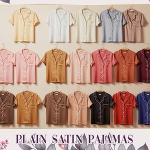 Può includere: Una collezione di pigiami in raso in vari colori, tra cui giallo, bianco, blu e rosa. Ogni top pigiama a maniche corte è esposto su una gruccia. Il testo "PLAIN SATIN PAJAMAS" è in basso nell'immagine.