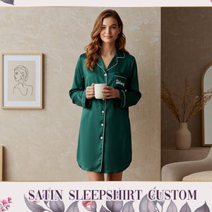 May include: A dark green satin sleepshirt with white trim and buttons. The sleepshirt has long sleeves and a personalized name embroidered on the chest. The model is holding a white mug. The text "SATIN SLEEPSHIRT CUSTOM" is at the bottom.
