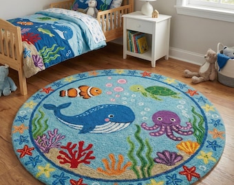 Op de afbeelding: Een ronde, lichtblauwe vloerkleed met een onderwaterthema. Het toont een blauwe walvis, een oranje clownvis, een groene zeeschildpad en een paarse octopus. Het kleed is omgeven door zeesterren en koraal.