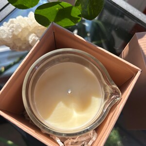 May include: A clear glass candle in a light brown box. The candle has a creamy white wax and a silver wick. The box is open, revealing the candle and shredded paper. Green leaves and a white crystal are in the background.