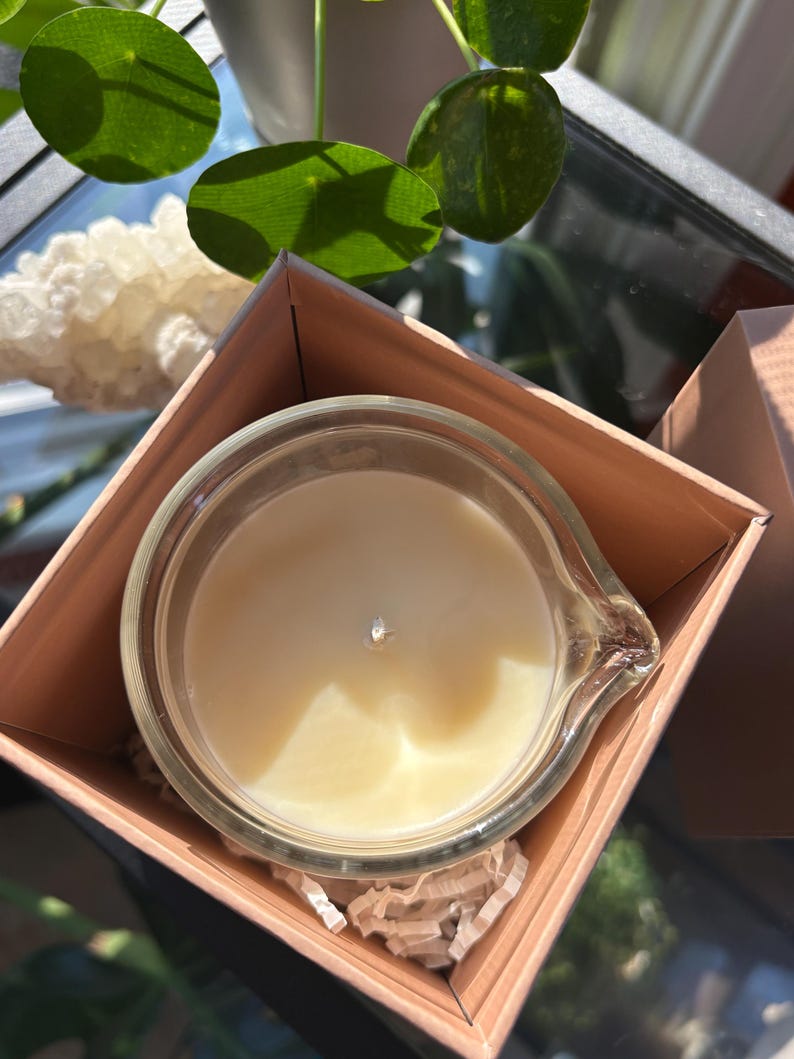 May include: A clear glass candle in a light brown box. The candle has a creamy white wax and a silver wick. The box is open, revealing the candle and shredded paper. Green leaves and a white crystal are in the background.