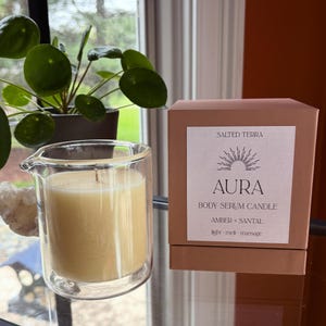 May include: Aura body serum candle in a clear glass container, with a light yellow wax and a wick. The candle is next to a brown box with the text "Aura Body Serum Candle Amber + Santal". A green plant is in the background.