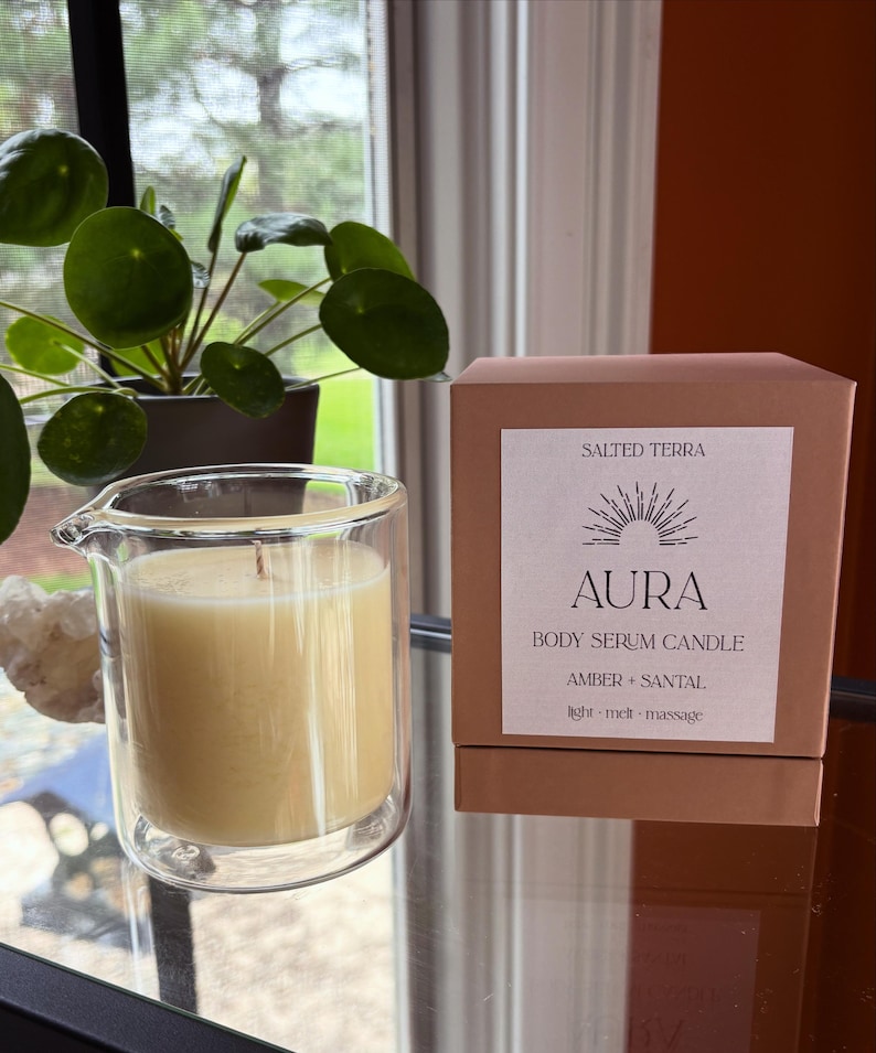 May include: Aura body serum candle in a clear glass container, with a light yellow wax and a wick. The candle is next to a brown box with the text "Aura Body Serum Candle Amber + Santal". A green plant is in the background.