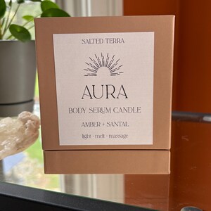 May include: A brown box with a white label featuring the text "SALTED TERRA" and "AURA BODY SERUM CANDLE." The label also includes the scent description "AMBER + SANTAL" and the words "light &middot; melt &middot; massage."