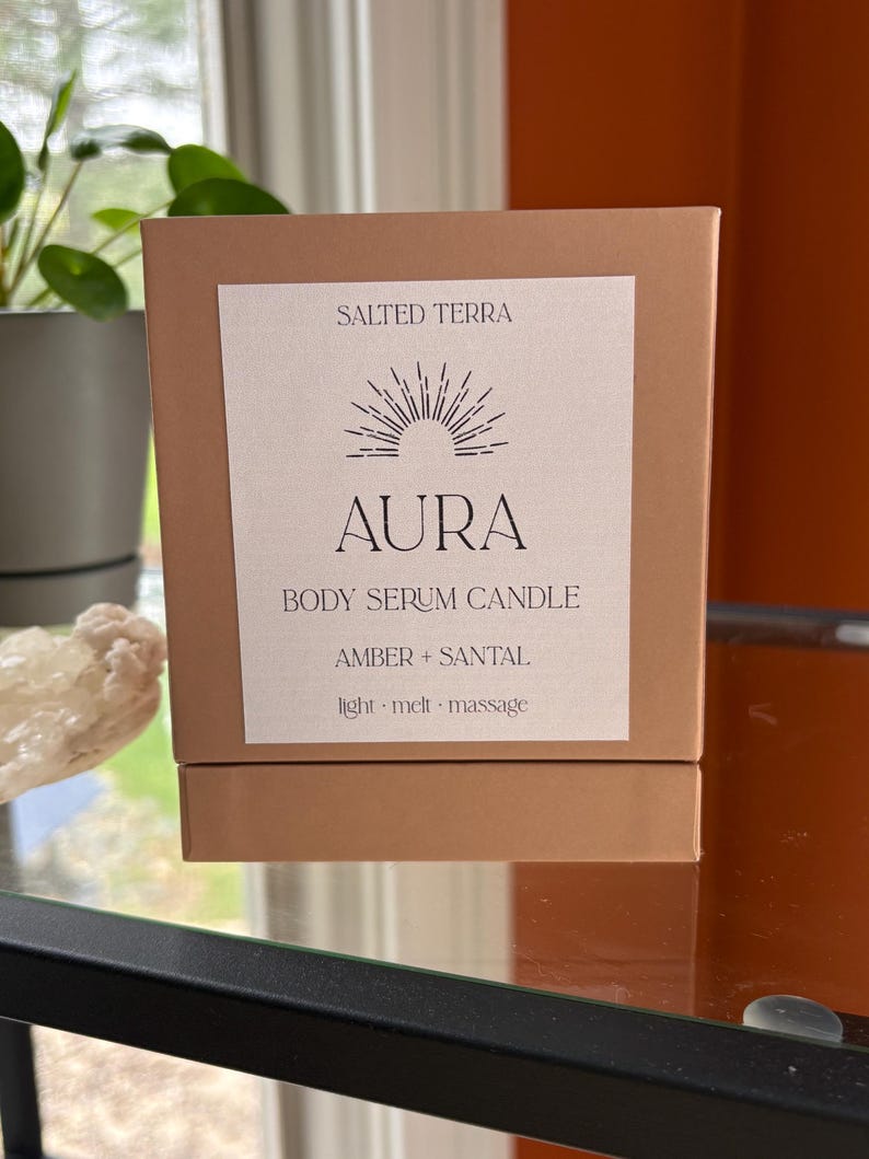 May include: A brown box with a white label featuring the text "SALTED TERRA" and "AURA BODY SERUM CANDLE." The label also includes the scent description "AMBER + SANTAL" and the words "light &middot; melt &middot; massage."