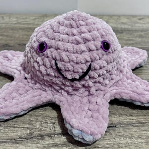 Peut inclure: Une pieuvre en peluche faite à la main, de couleur lavande douce. La pieuvre a un visage souriant avec des détails cousus en noir et des yeux violets. Les tentacules sont étalés et les bords ont une teinte bleu clair.