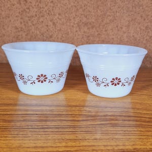 May include: Two white cups with a reddish-brown floral pattern. The cups are on a wooden surface with a brown background. The cups have a slightly flared shape. The cups are vintage-style.