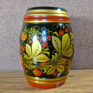 May include: A hand-painted, barrel-shaped wooden container. The black body is decorated with gold leaves, red berries, and orange flowers. The top and bottom rims are red and gold. A decorative piece.