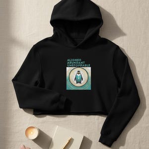 May include: Black cropped hoodie with a hood. The front features a square graphic with a penguin illustration and the words "ALIGNED ABUNDANT UNSTOPPABLE". A candle, notebook, pen, and crystals are in the foreground.