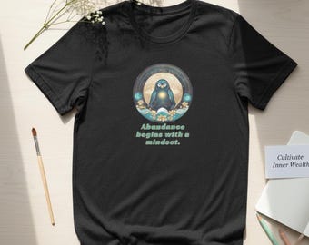 May include: A black short-sleeved t-shirt featuring a circular graphic of a blue penguin centered on the chest. The graphic includes a glowing light, flowers, and the text "Abundance begins with a mindset." in a light green, sans-serif font.