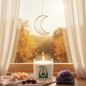 May include: A lit candle in a clear glass jar with the text "Aligned Abundant Unstoppable" and a penguin graphic. Beside the candle are a small bowl of candy corn, three brown nuts, and a purple amethyst crystal. A crescent moon decoration hangs in front of a window.