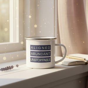 May include: A white enamel mug with a silver rim, featuring the words "ALIGNED," "ABUNDANT," and "UNSTOPPABLE" in navy blue. The mug sits on a windowsill next to a sprig of lavender and an open book.