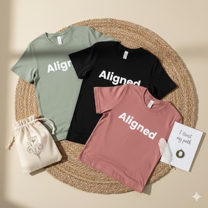 Puede incluir: Tres camisetas de manga corta en verde salvia, negro y rosa empolvado, cada una con la palabra "Aligned" impresa en blanco. También hay una pequeña bolsa con cordón con un diseño floral y una tarjeta con el texto "I trust my path".