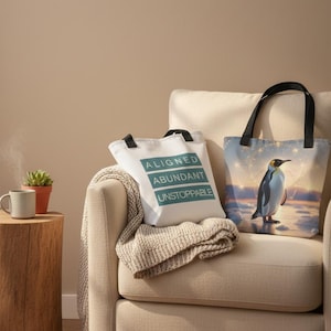 May include: Two tote bags rest on a beige armchair. One is white with the words "ALIGNED, ABUNDANT, UNSTOPPABLE" in teal. The other features a penguin illustration against a sunset backdrop. A small succulent and a mug sit on a wooden side table.
