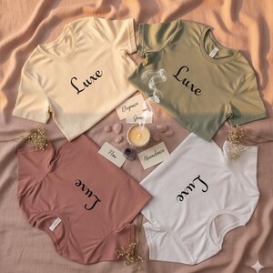 May include: Four short-sleeved t-shirts in cream, olive green, dusty rose, and white, each with the word "Luxe" in black script. The shirts are arranged on a pink fabric with a candle, crystals, and small cards.