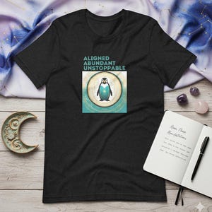 May include: Dark gray t-shirt with a graphic of a penguin inside a circle, with the words "ALIGNED ABUNDANT UNSTOPPABLE" above it. The shirt is on a white surface with a notebook, pen, and crystals.