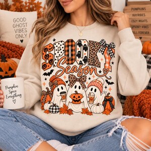 Puede incluir: Una sudadera beige de cuello redondo con un estampado gráfico que incluye el texto SPOOKY Season. El diseño muestra tres fantasmas, calabazas, hojas de otoño, murciélagos y estrellas. Los fantasmas llevan sombreros y lazos con estampados.