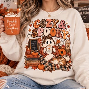 Puede incluir: Una sudadera color crema con un estampado gráfico que incluye el texto "COZY & SPOOKY". El diseño muestra un fantasma con un lazo de leopardo, un gato durmiendo, calabazas, una pila de libros y una taza con "PUMPKIN SPICE EVERYTHING".