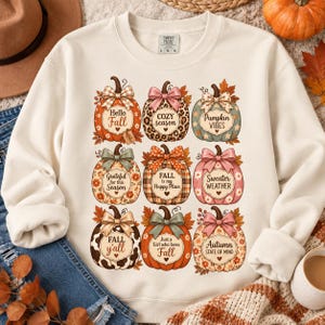 Puede incluir: Una sudadera de cuello redondo color crema con un estampado gráfico de nueve calabazas dispuestas en cuadrícula. Cada calabaza tiene un patrón único, un lazo y texto que incluye Hello Fall, Cozy Season, Pumpkin Vibes, Grateful, Fall is my Happy Place, Sweater Weather, Fall y'all, Just a Girl who loves Fall y Autumn State of Mind.