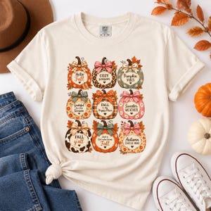 Puede incluir: Una camiseta color crema con una cuadrícula de nueve calabazas ilustradas, cada una con un patrón y un lazo únicos. Las calabazas incluyen textos como Hello Fall, Cozy season, Pumpkin VIBES, Grateful for this Season, FALL is my Happy Place.