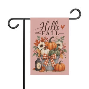 Puede incluir: Una bandera de jardín rectangular en un soporte de metal negro con fondo rosa empolvado, el texto Hello FALL y un corazón. El diseño incluye un cubo de metal lleno de calabazas, flores blancas y hojas de otoño, además de un lazo a cuadros.