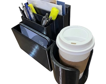 Puede incluir: Organizador de escritorio negro con compartimentos para smartphone, bolígrafos y correo. Una taza de café con tapa blanca y funda marrón se encuentra en un portavasos incorporado. Hecho de plástico negro.